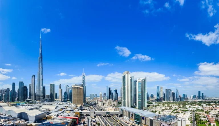 Wide panoramic view of the Dubai skyline featuring the Burj Khalifa, modern skyscrapers, and city highways under a blue sky.
