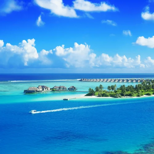 Luxurious overwater bungalows and a verdant tropical island encircled by the vivid blue waters of the Maldives are seen from above.