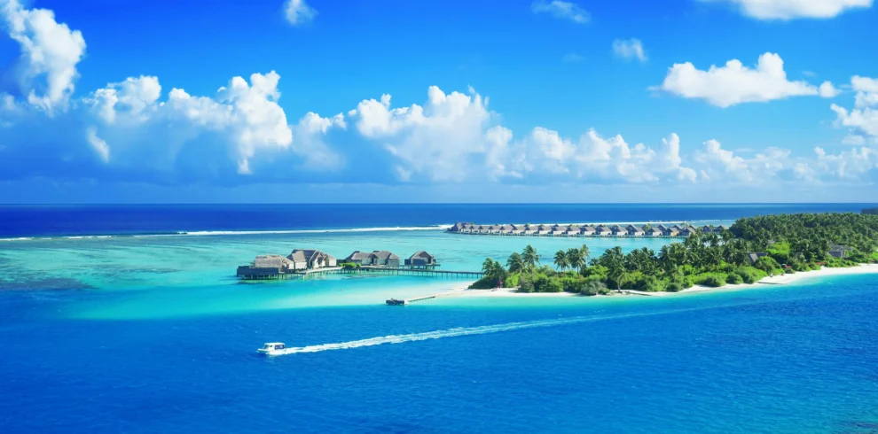 Luxurious overwater bungalows and a verdant tropical island encircled by the vivid blue waters of the Maldives are seen from above.