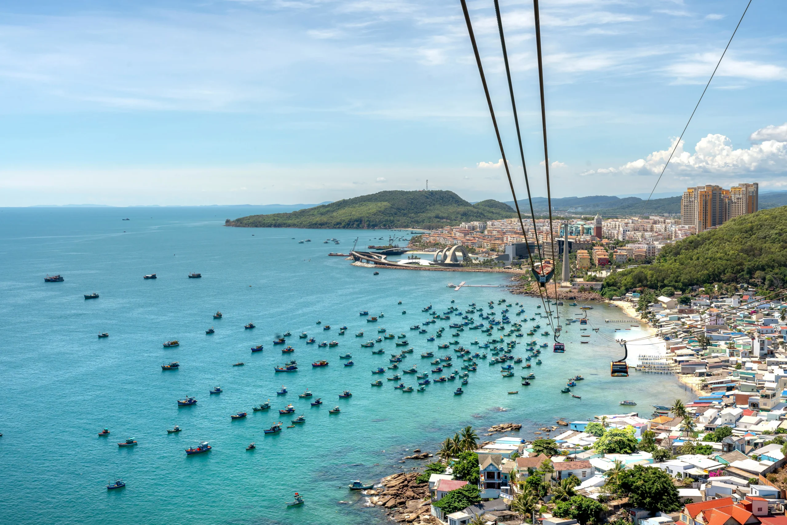Aerial view of Phu Quoc cable car over turquoise water with many blue fishing boats and coastal town in Vietnam.