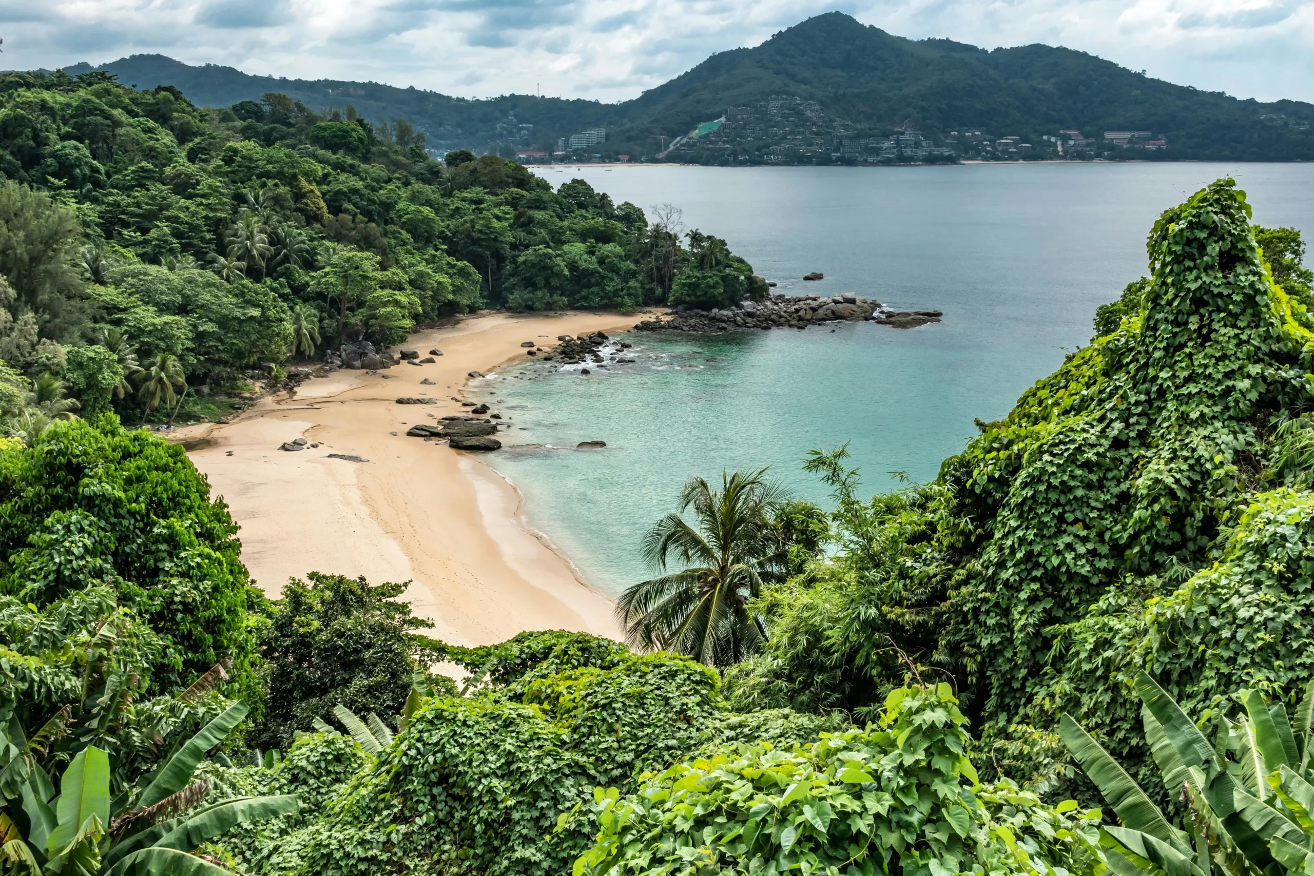 High-angle view of a hidden sandy beach in Phuket, Thailand, surrounded by lush tropical jungle and calm turquoise water.