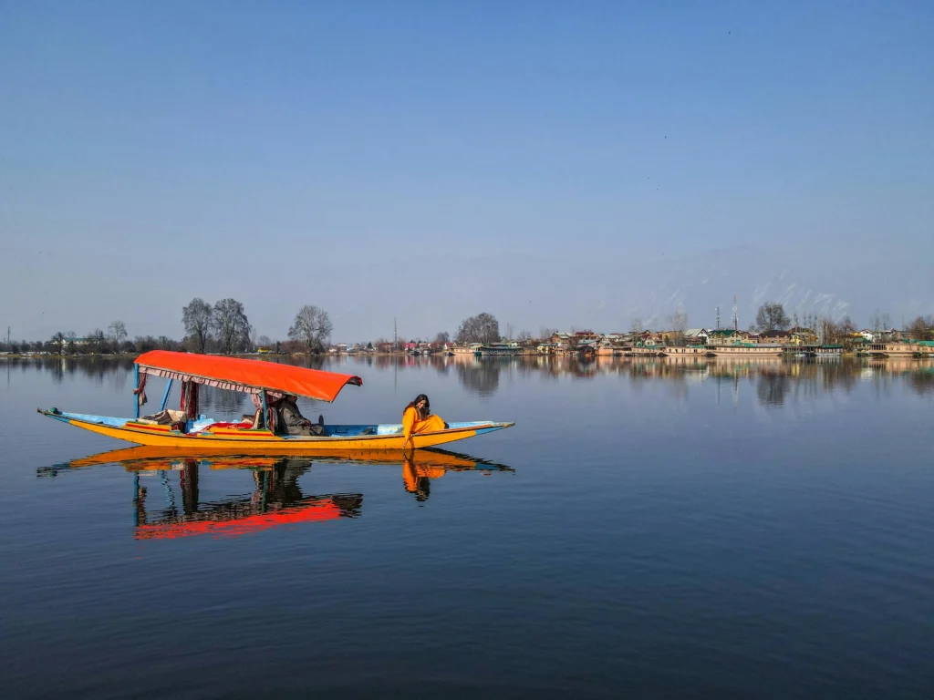 A colorful traditional Shikara boat with an orange canopy gliding on the calm, reflective waters of Dal Lake in Kashmir.