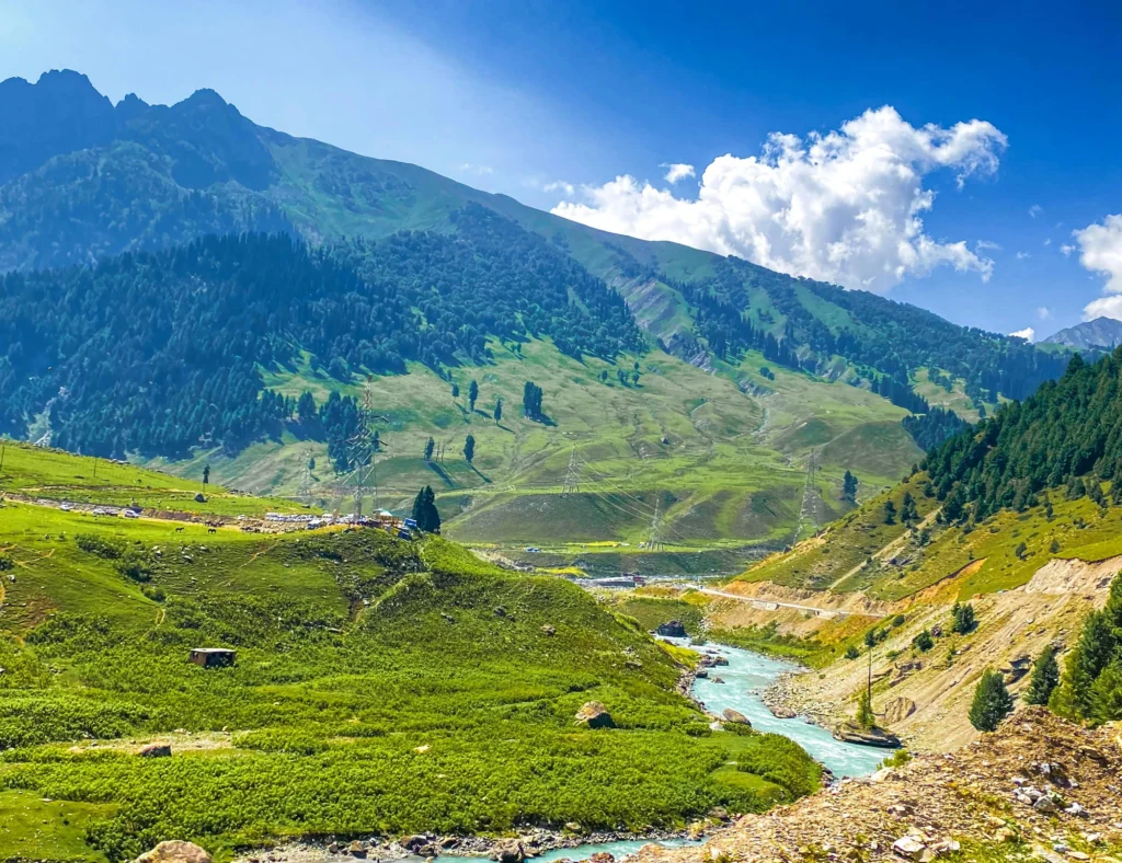 A blue river flowing through green mountains in Sonamarg, Kashmir, under a bright sunny sky.