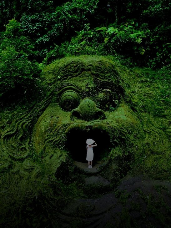 A tourist standing inside a massive moss-covered stone face structure in a Bali jungle.